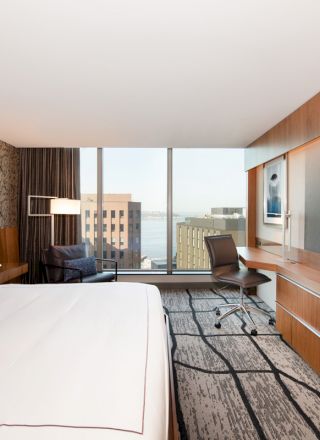 The image shows a modern hotel room with a large bed, a seating area by the window, a desk, a chair, a flat-screen TV, and a view of city buildings.