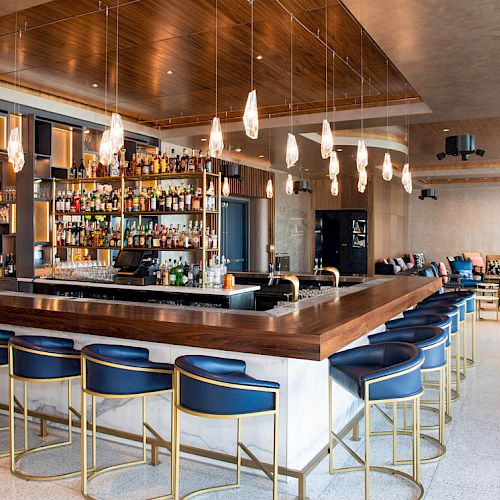 The image shows a modern bar with a sleek design, featuring blue bar stools with gold frames, hanging lights, and a well-stocked bar counter.