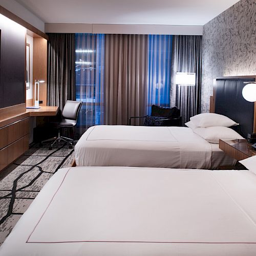 A modern hotel room features two neatly made beds, a TV, a desk with a chair, and a large window with curtains.