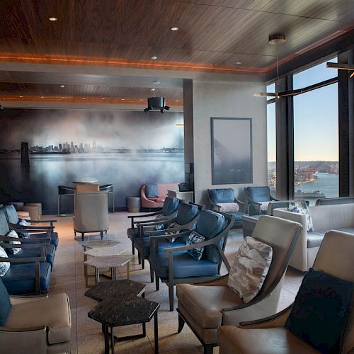 A modern lounge area with multiple armchairs and tables, a wall mural, large windows offering a waterfront view, and contemporary lighting fixtures.