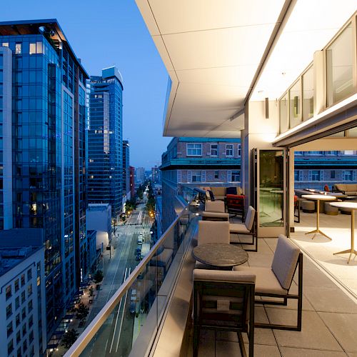 A modern balcony overlooks a cityscape at dusk, with comfortable seating and tables. The scene includes tall buildings and an urban street below.
