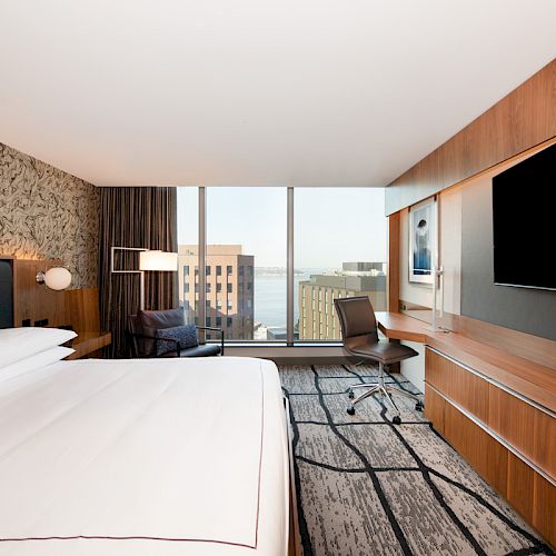 This image shows a modern hotel room with a large bed, a desk, a chair, a TV on the wall, and a window with a city view.