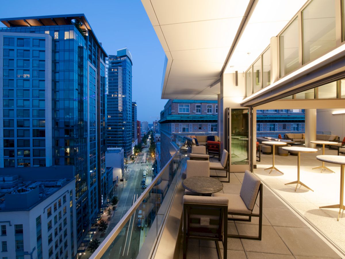 A modern rooftop balcony at dusk with tables and chairs, overlooking a city street lined with tall glass buildings and illuminated windows.