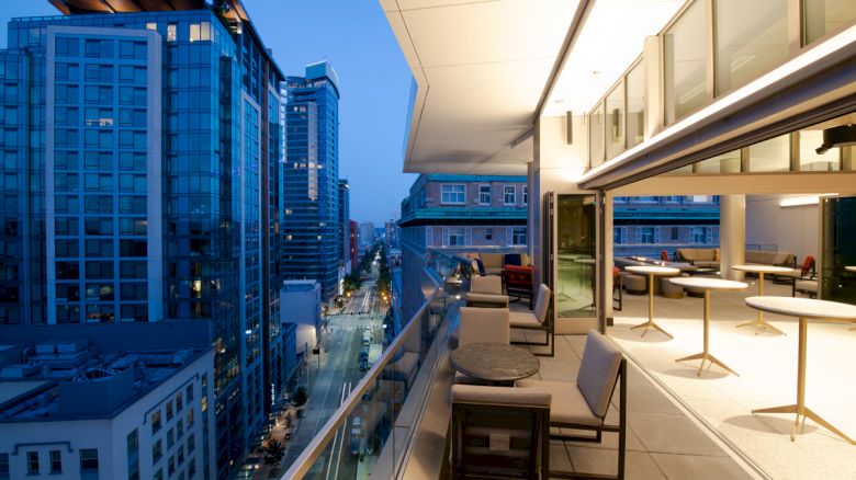A modern rooftop balcony at dusk with tables and chairs, overlooking a city street lined with tall glass buildings and illuminated windows.