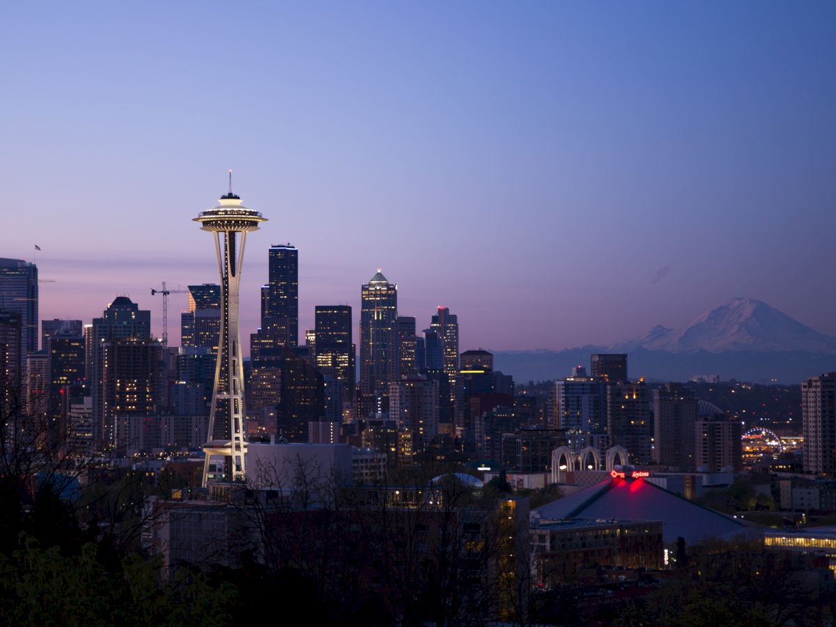 The image shows the Seattle skyline at dusk, featuring the Space Needle prominently. In the background, Mount Rainier is visible.