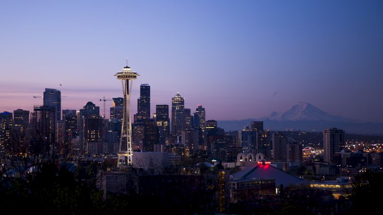 The image shows the Seattle skyline at dusk, featuring the Space Needle prominently. In the background, Mount Rainier is visible.