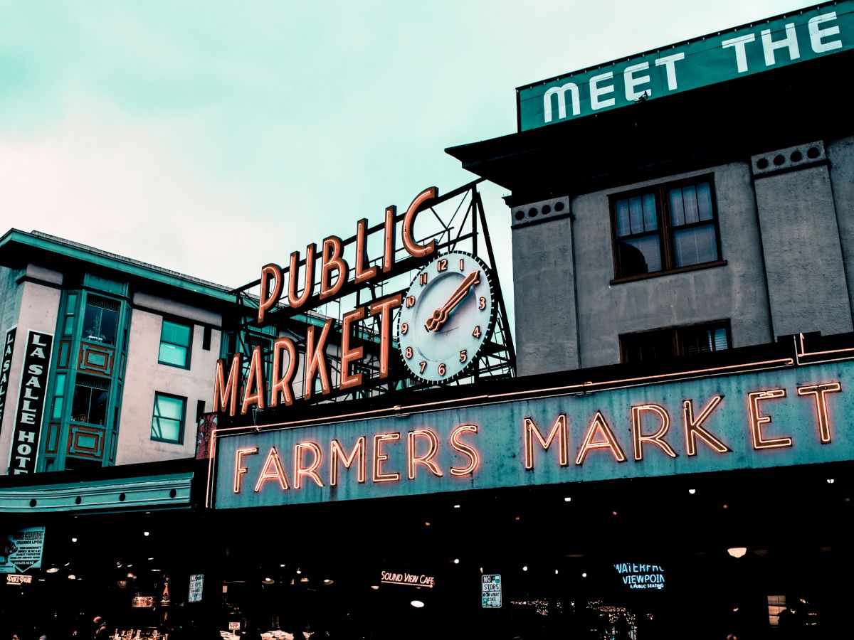 The image shows the iconic neon sign of a public market and farmers market in an urban setting with buildings and a clock.