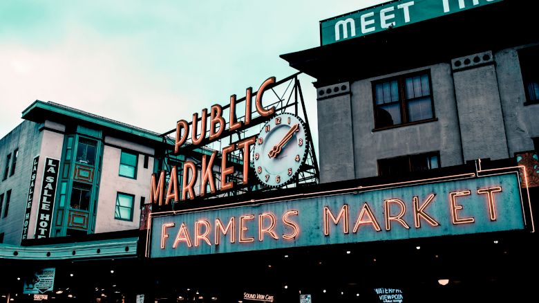 The image shows the iconic neon sign of a public market and farmers market in an urban setting with buildings and a clock.