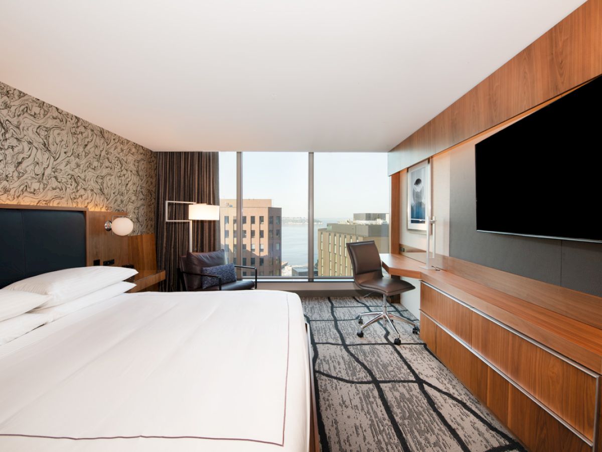 A modern hotel room features a large bed, a TV, a desk, a floor-to-ceiling window with a city view, and artwork on the walls.