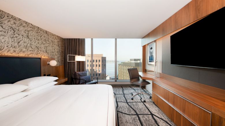 A modern hotel room features a large bed, a TV, a desk, a floor-to-ceiling window with a city view, and artwork on the walls.