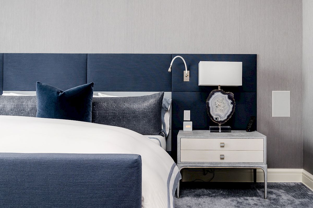 A modern bedroom features a blue upholstered headboard, white nightstand, table lamp, and a neatly made bed with blue accents, ending the sentence.