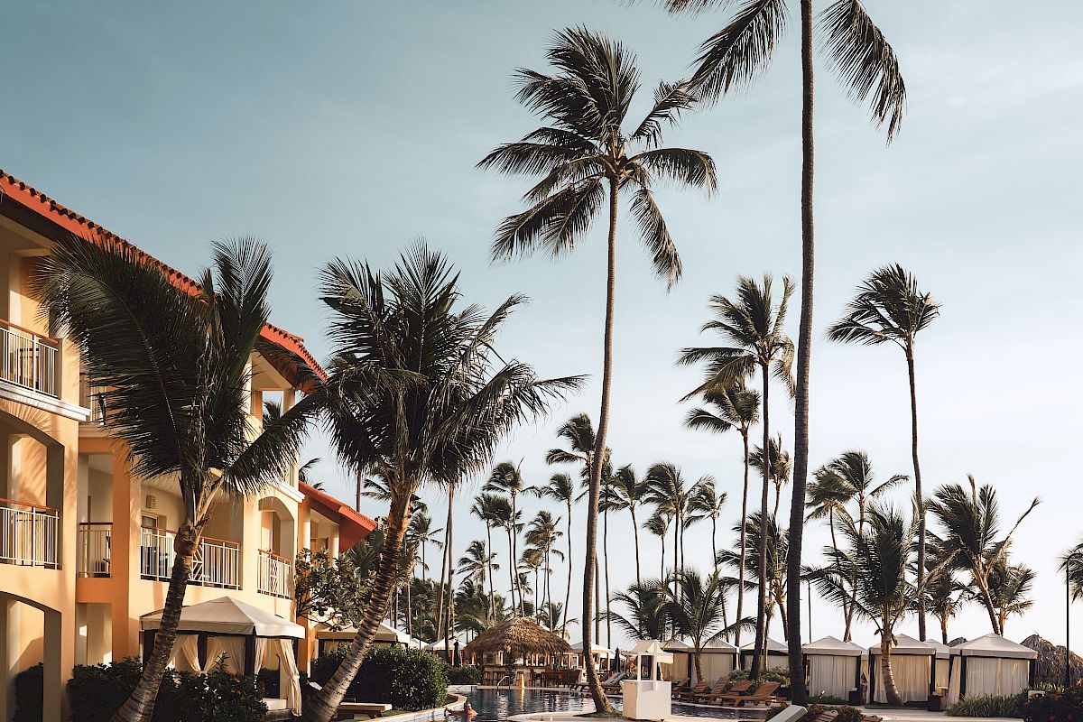 A serene pool area with lounge chairs and tall palm trees, located next to a multi-story building with balconies, under a clear sky.