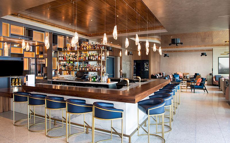 The image shows a modern bar with a wooden countertop, high chairs, a well-stocked back bar, hanging lights, and a seating area in the background.