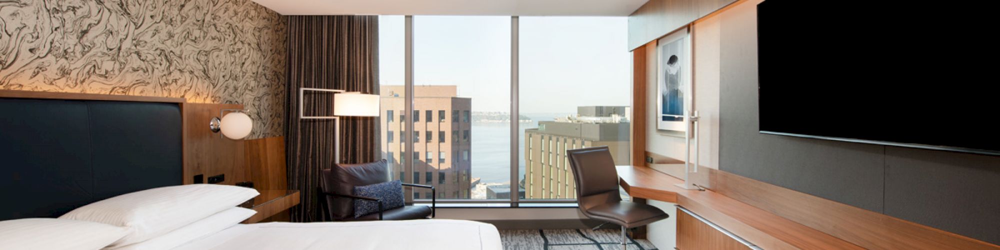A modern hotel room with a large bed, desk, chair, TV, and a window with a city view is depicted in the image.