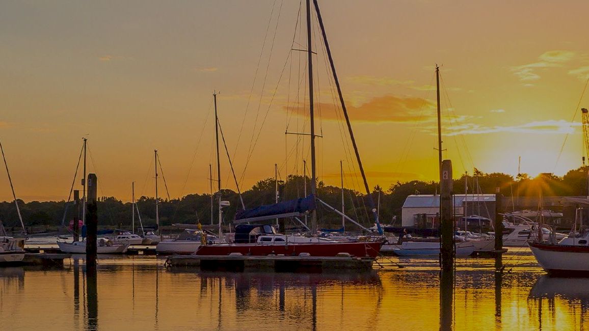 A serene marina scene at sunset with several sailboats docked on calm waters, casting reflections.