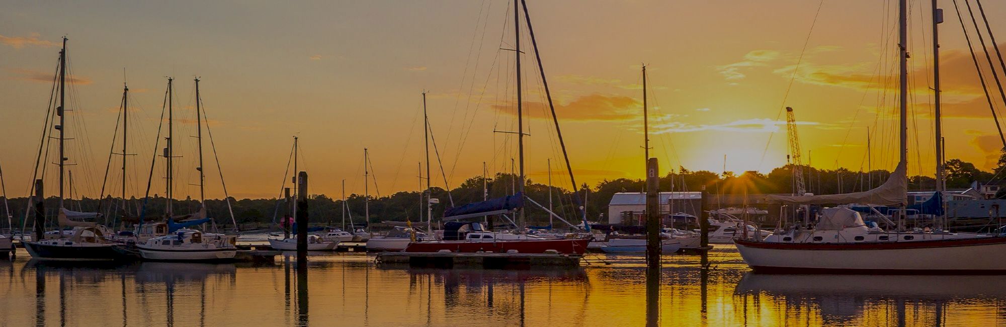 A serene marina scene at sunset with several sailboats docked on calm waters, casting reflections.