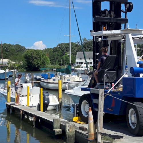 A dockside marina scene with boats tied up, a worker loading gear, and a truck-mounted lift crane under a clear blue sky.