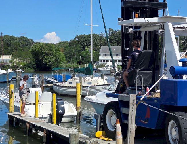 A dockside marina scene with boats tied up, a worker loading gear, and a truck-mounted lift crane under a clear blue sky.