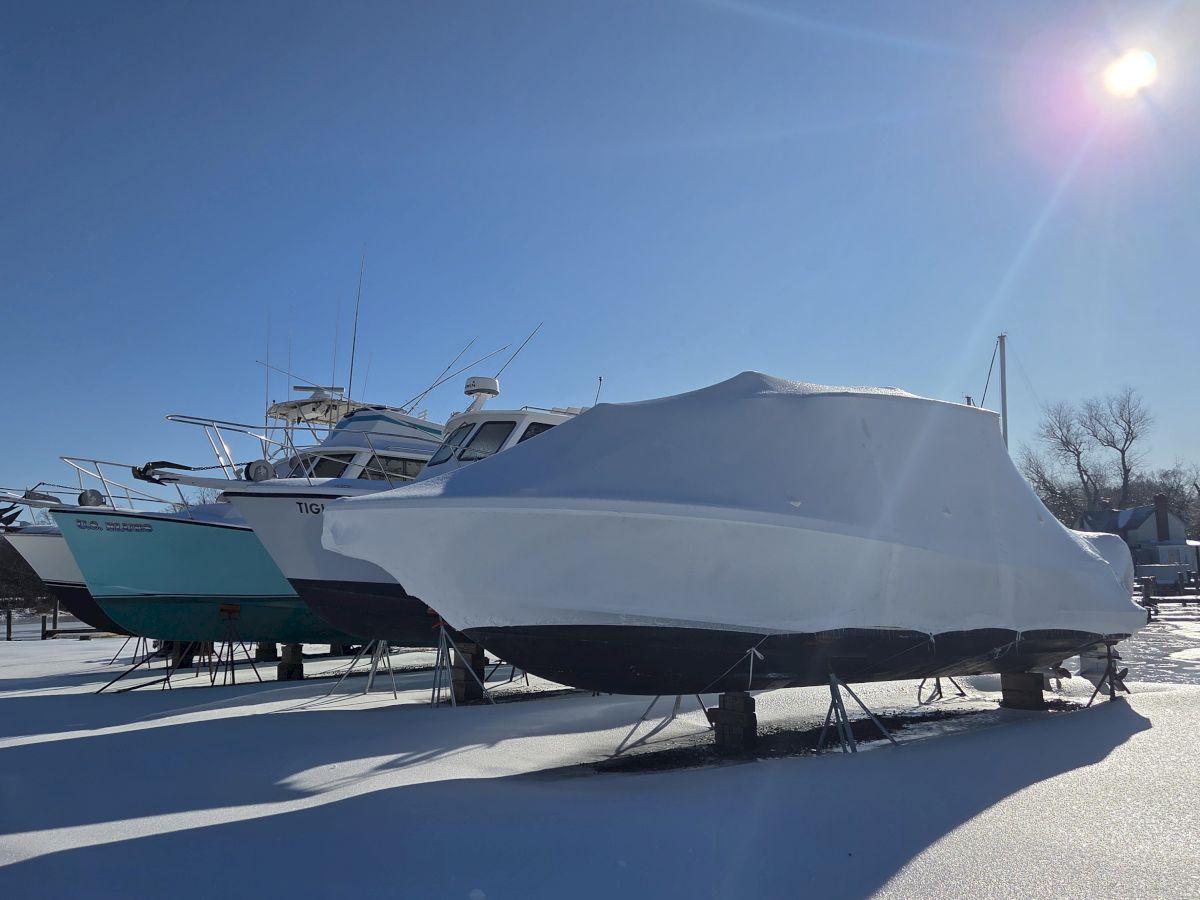 Snowy marina yard with a white-and-blue motorboat/cabin cruiser hauled on stands, under bright sun and lens flare.