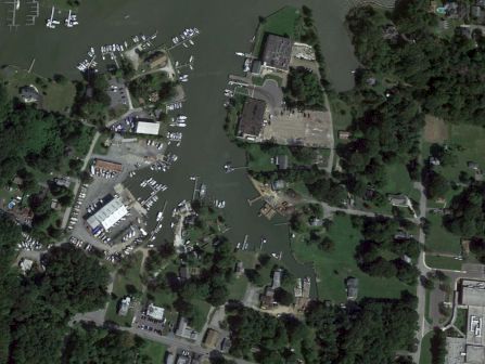 An aerial view of a marina with boats docked, surrounded by trees, buildings, and open areas, likely in a coastal or lakeside town.