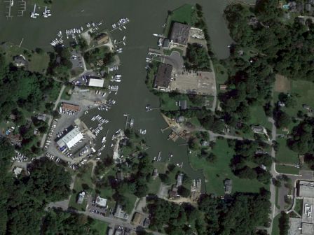 An aerial view of a marina with boats docked, surrounded by trees, buildings, and open areas, likely in a coastal or lakeside town.