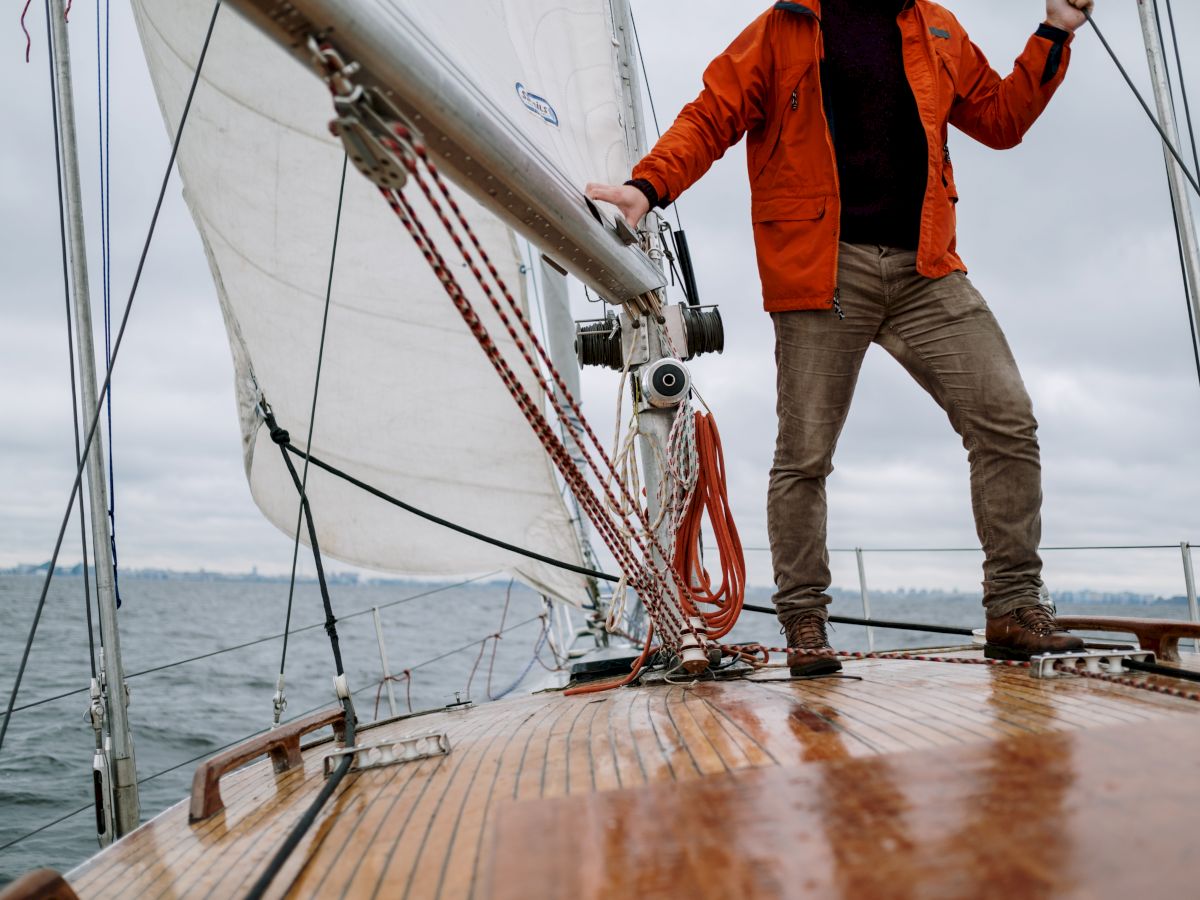 A person stands on the deck of a sailboat, holding onto the mast, with the sails partially opened and the sea in the background.
