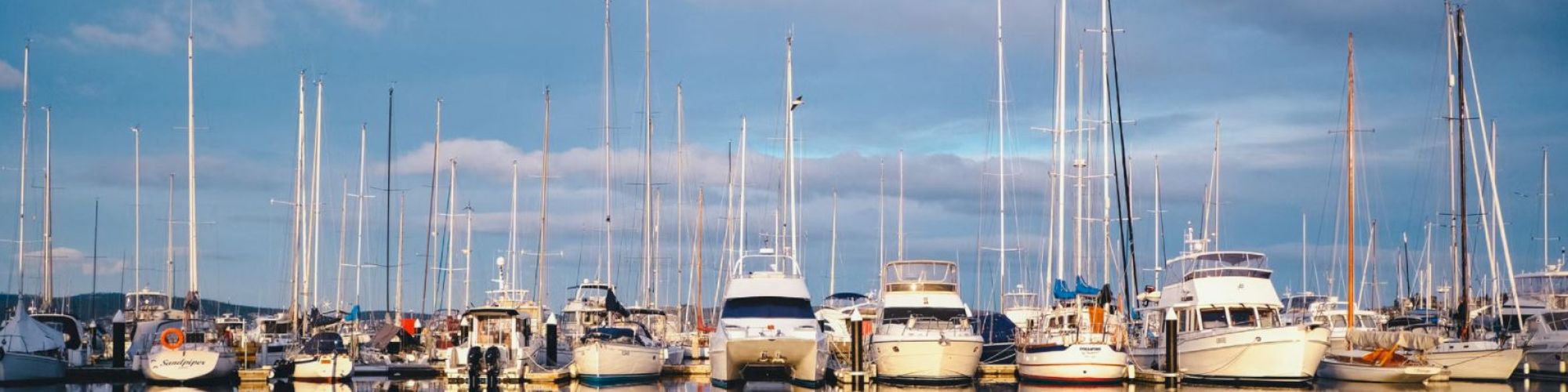 A marina with numerous yachts and sailboats docked, reflecting on the calm water under a partly cloudy sky.