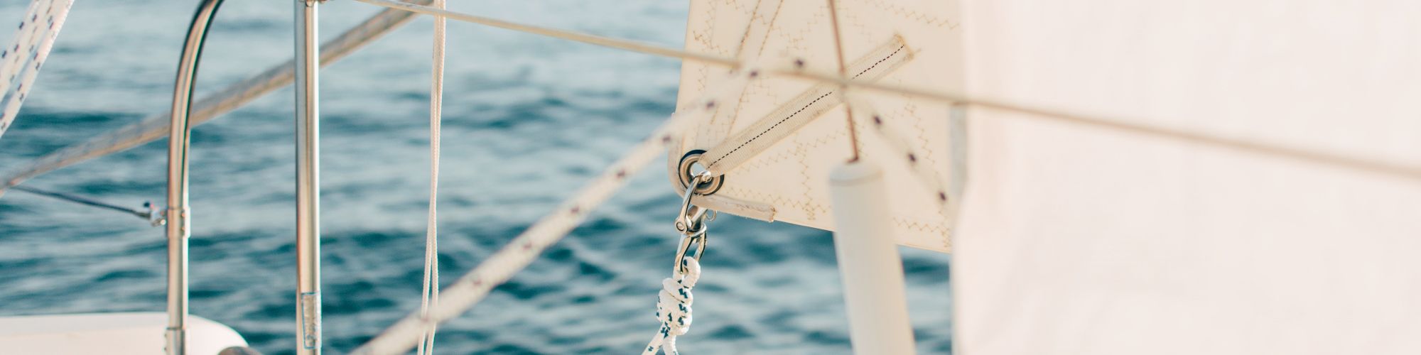A close-up view of a sailboat's deck and rigging, showing parts of the sail and metallic fixtures, with calm water in the background.