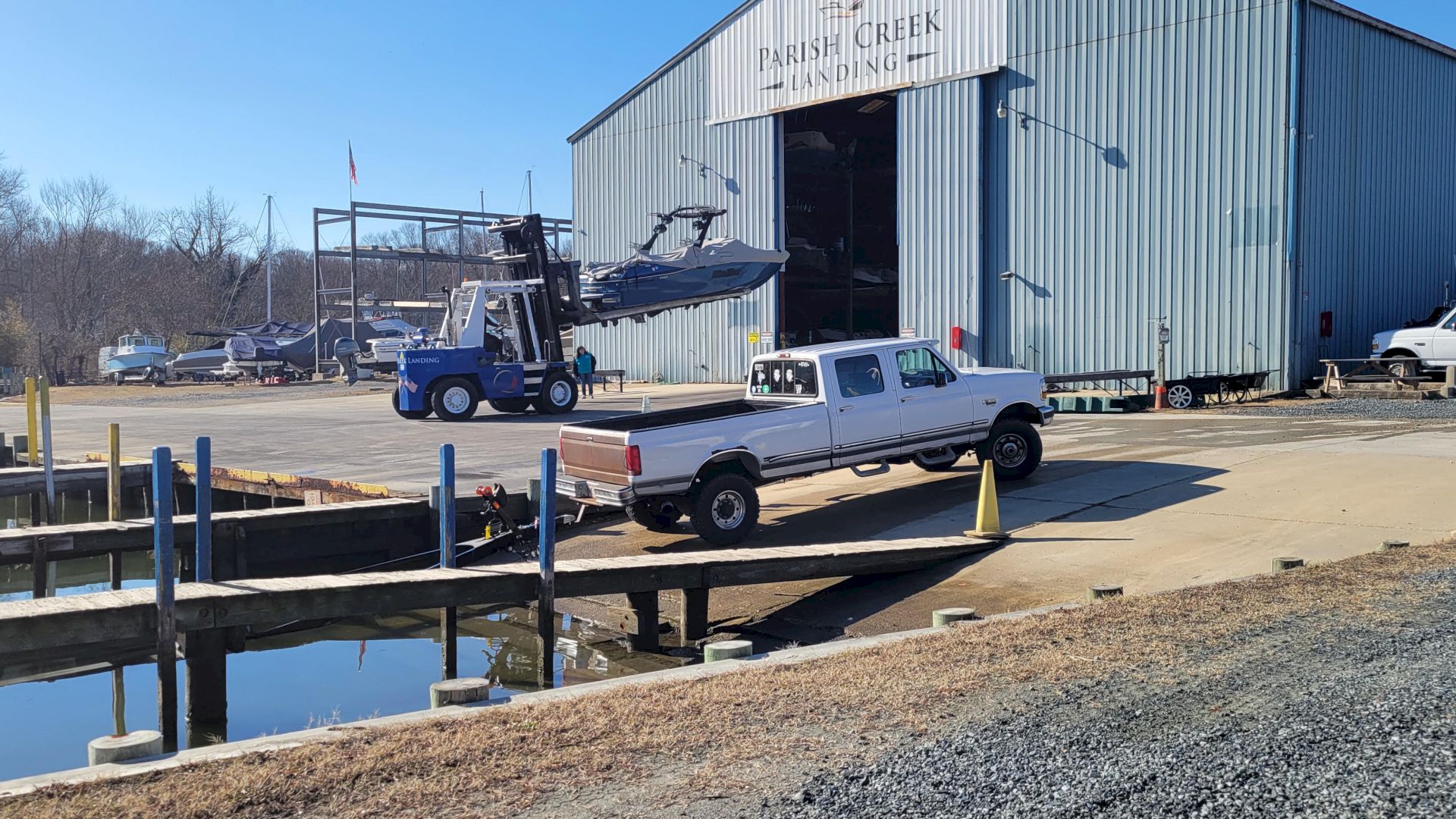 A forklift lifts a boat for loading beside a dock while a pickup truck waits near a large &ldquo;Parish Greek Landing&rdquo; warehouse.