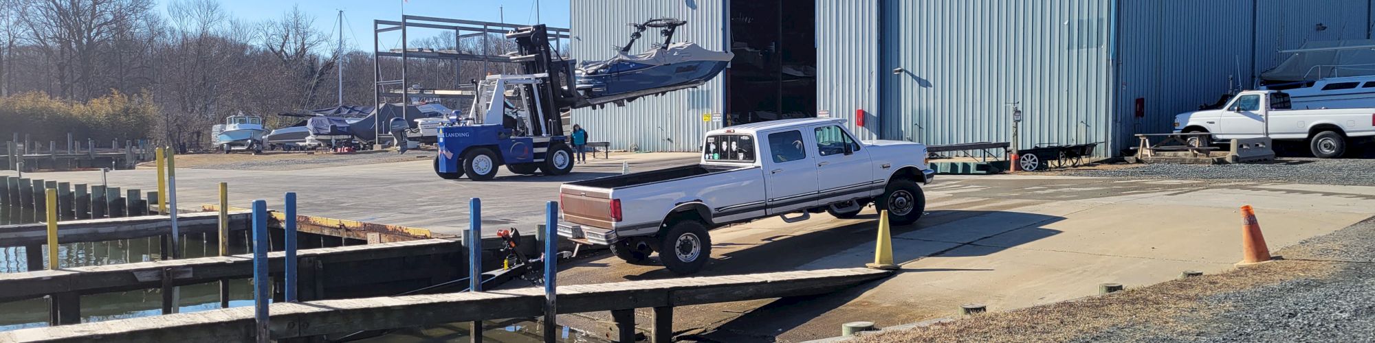 A forklift lifts a boat for loading beside a dock while a pickup truck waits near a large &ldquo;Parish Greek Landing&rdquo; warehouse.