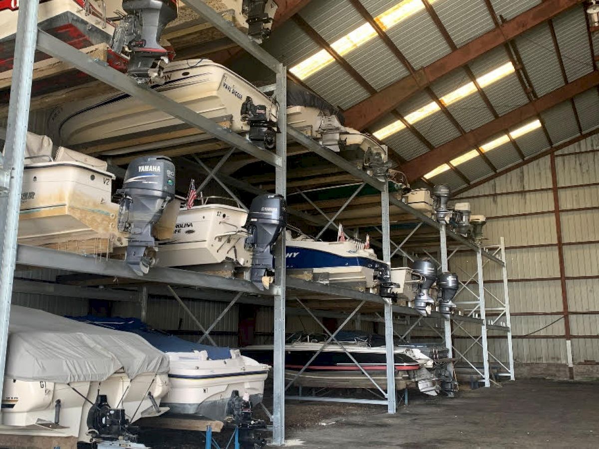 The image shows boats stored on racks inside a large warehouse with a high, slanted roof and metal structure.